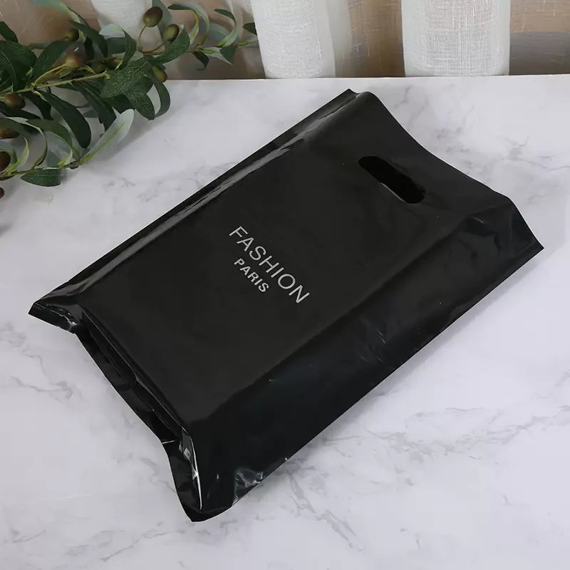 Black glossy plastic retail bag printed with white "FASHION PARIS" logo, displayed on a marble surface with decorative greenery.
