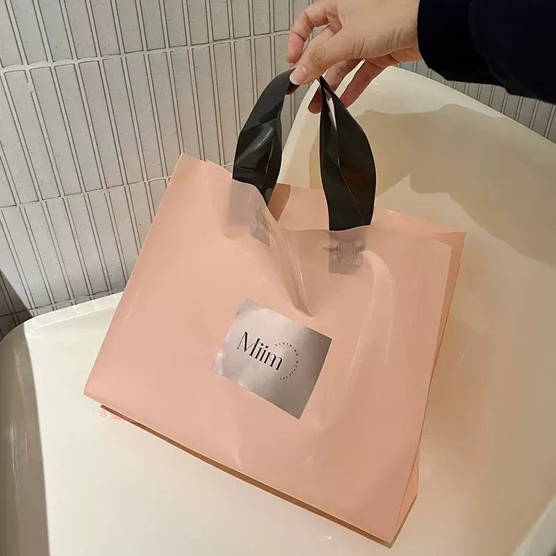 Pink reusable tote bag (black handles) with a metallic "Miim" label, held by a hand against a tiled background.