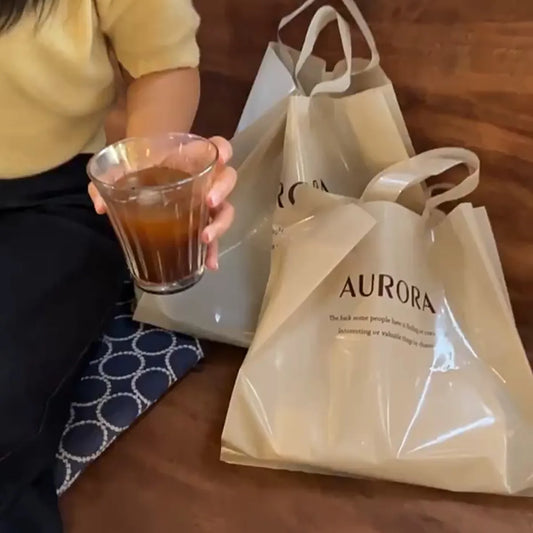 Beige reusable shopping bags printed with "AURORA" and a text quote, placed next to a person holding a glass of iced drink (casual setting)