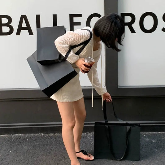 Woman holding multiple black reusable shopping bags (and an iced drink) outside a retail store, showcasing post-shopping use of eco-friendly bags.