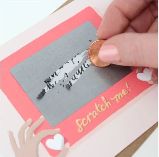 Handmade scratch-off card craft in progress: a person uses a penny to scratch the gray coating on a pink-themed card, revealing hidden text (labeled "scratch me!").