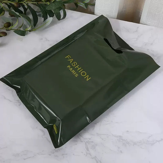 Olive green plastic retail bag printed with "FASHION PARIS" (glossy finish), displayed on a marble surface with decorative greenery.