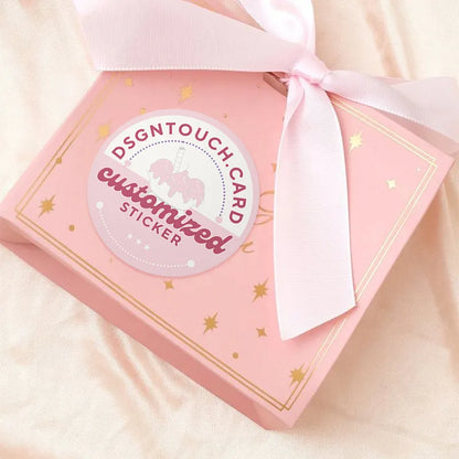 Customized "DSGNTouch" sticker for gift packaging: pink & gold design (with unicorn graphic) applied to a star-accented pink gift box, tied with a satin ribbon.